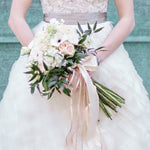 Closeup of a bride holding a bridal bouquet featuring white and soft pink flowers with a soft pink ribbon tied around the stems.